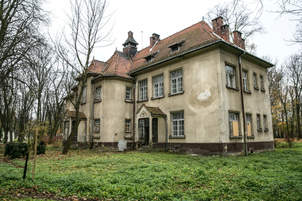 Budynek w Kobierzynie Stara, zaniedbana willa o dwuspadowym dachu z czerwonej dachówki, otoczona drzewami bez liści oraz zarośniętym trawnikiem, widoczne zniszczenia elewacji i zabite deskami okna.