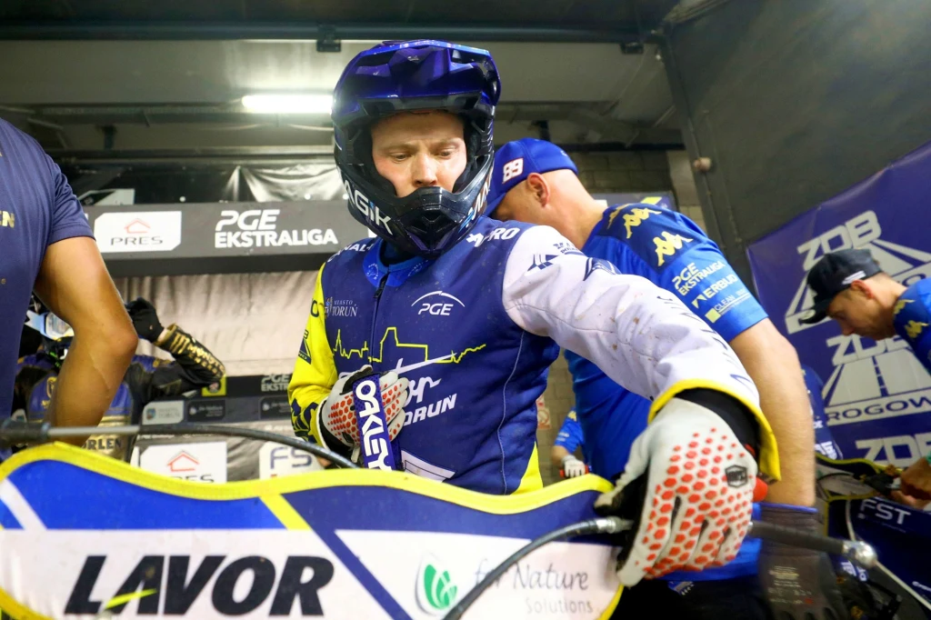 Emil Sajfutdinow w jednej z gonitw przeszedł z czwartej na pierwszą pozycję Zawodnik żużlowy w kolorowym kombinezonie i kasku przygotowuje motocykl do wyścigu w obecności zespołu technicznego, w tle widoczne banery sponsorskie i logotypy ligi.