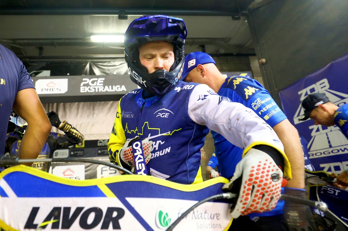 Zawodnik żużlowy w kolorowym kombinezonie i kasku przygotowuje motocykl do wyścigu w obecności zespołu technicznego, w tle widoczne banery sponsorskie i logotypy ligi.