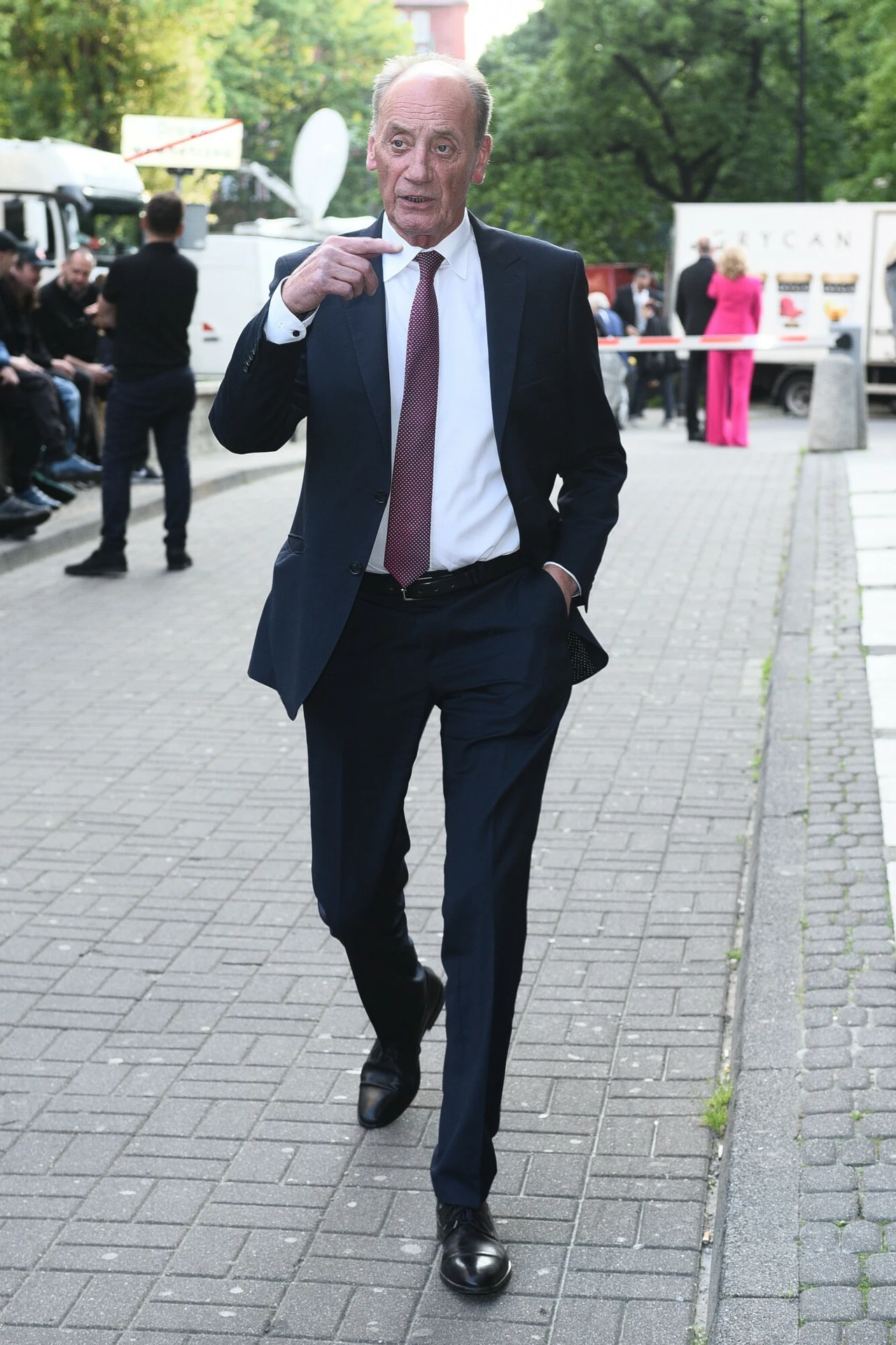 Dariusz Szpakowski Elegancki mężczyzna w ciemnym garniturze i bordowym krawacie idzie pewnym krokiem chodnikiem, z jedną ręką w kieszeni, na tle ludzi czekających przy ulicznych pojazdach oraz zieleni w tle.