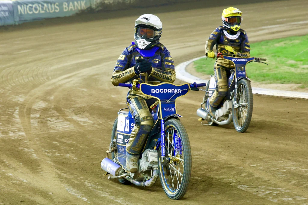 Dwóch żużlowców w pełnym rynsztunku sportowym porusza się po torze żużlowym, motocykle oznaczone logotypami sponsorów, za zawodnikami widoczne zakręty toru oraz fragmenty bandy z reklamami.