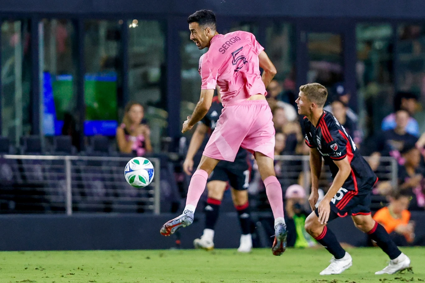 Sergio Busquets Dwóch piłkarzy w trakcie akcji na boisku podczas meczu piłki nożnej, zawodnik w różowej koszulce wykonuje zagranie głową, a zawodnik w czarnej koszulce obserwuje sytuację.
