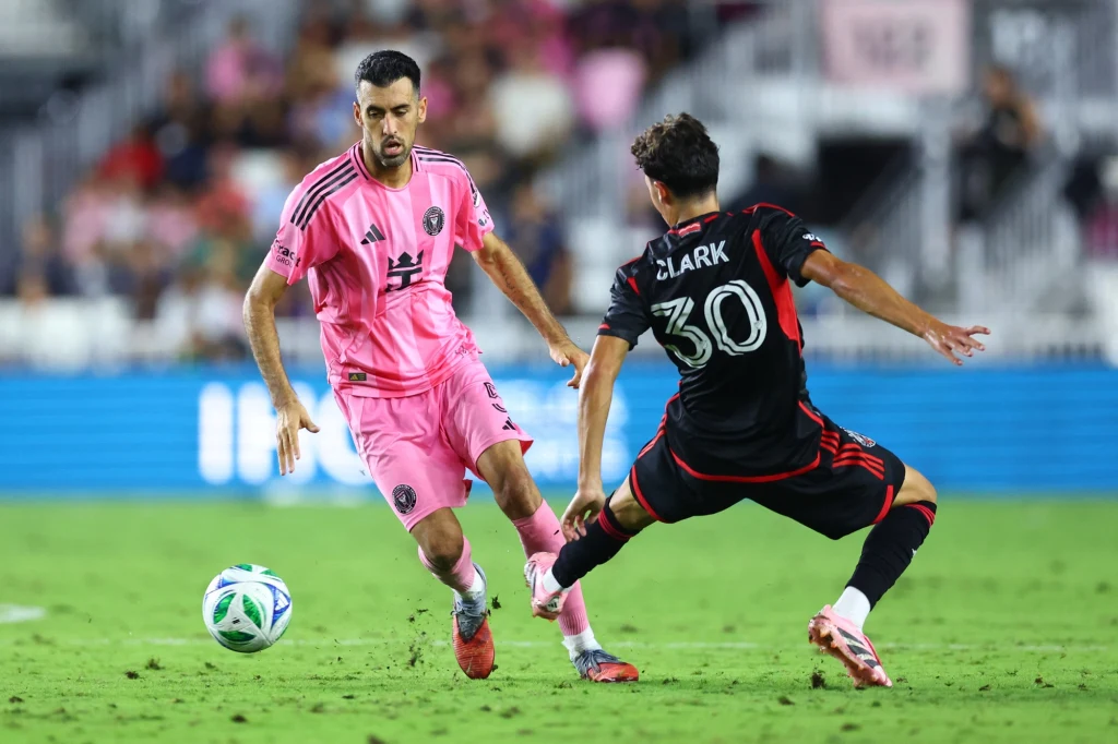 Sergio Busquets Dwóch piłkarzy rywalizuje o piłkę na murawie stadionu, jeden ubrany w różowy strój, drugi w czarny z numerem 30 na plecach, obaj są skupieni na grze i dynamicznie zaangażowani w akcję.