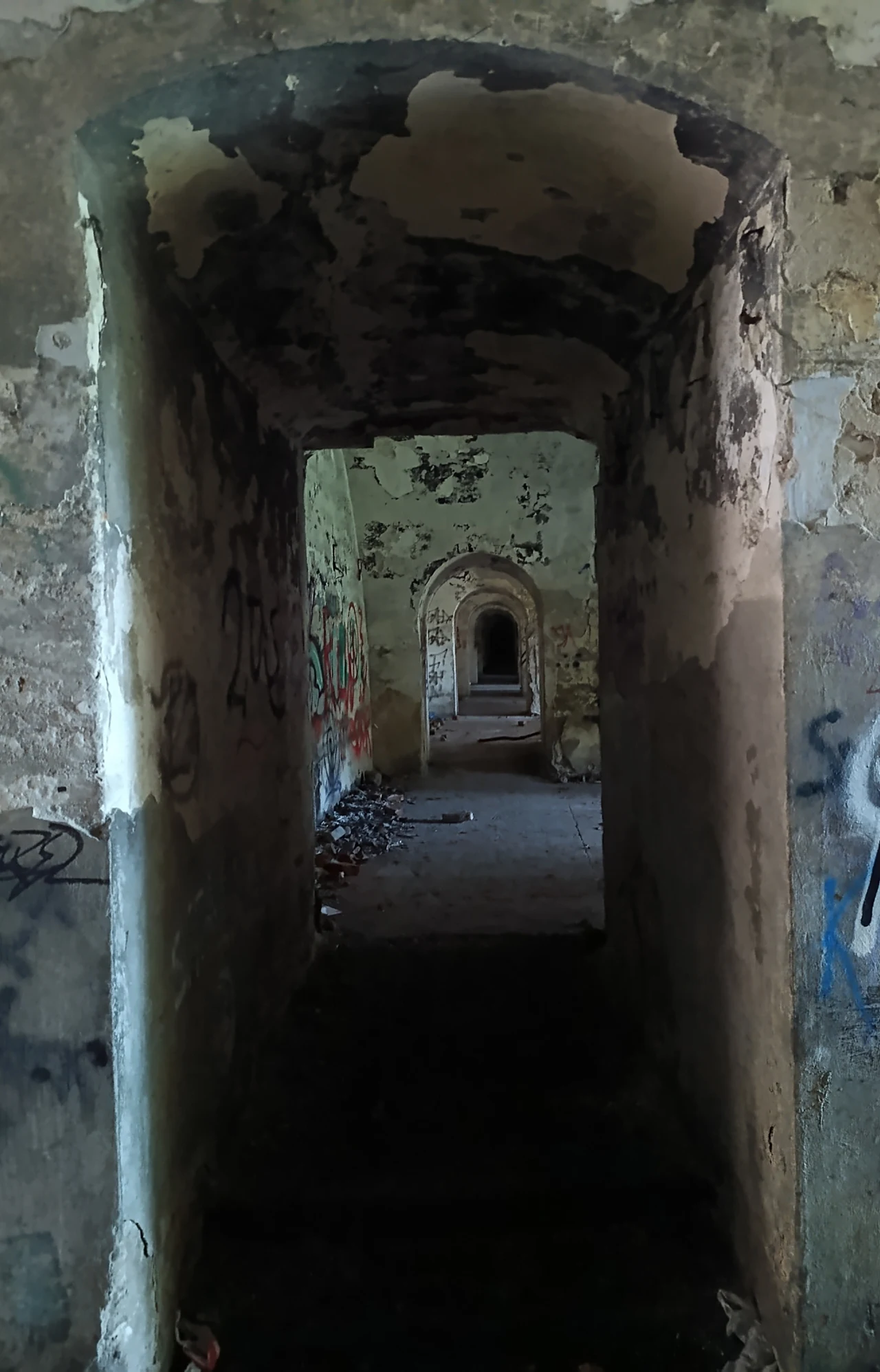 Korytarz w Forcie 50 Prokocim Zniszczone, opuszczone korytarze w starym budynku z widocznymi śladami wandalizmu i graffiti na ścianach, słabe oświetlenie, surowa atmosfera.