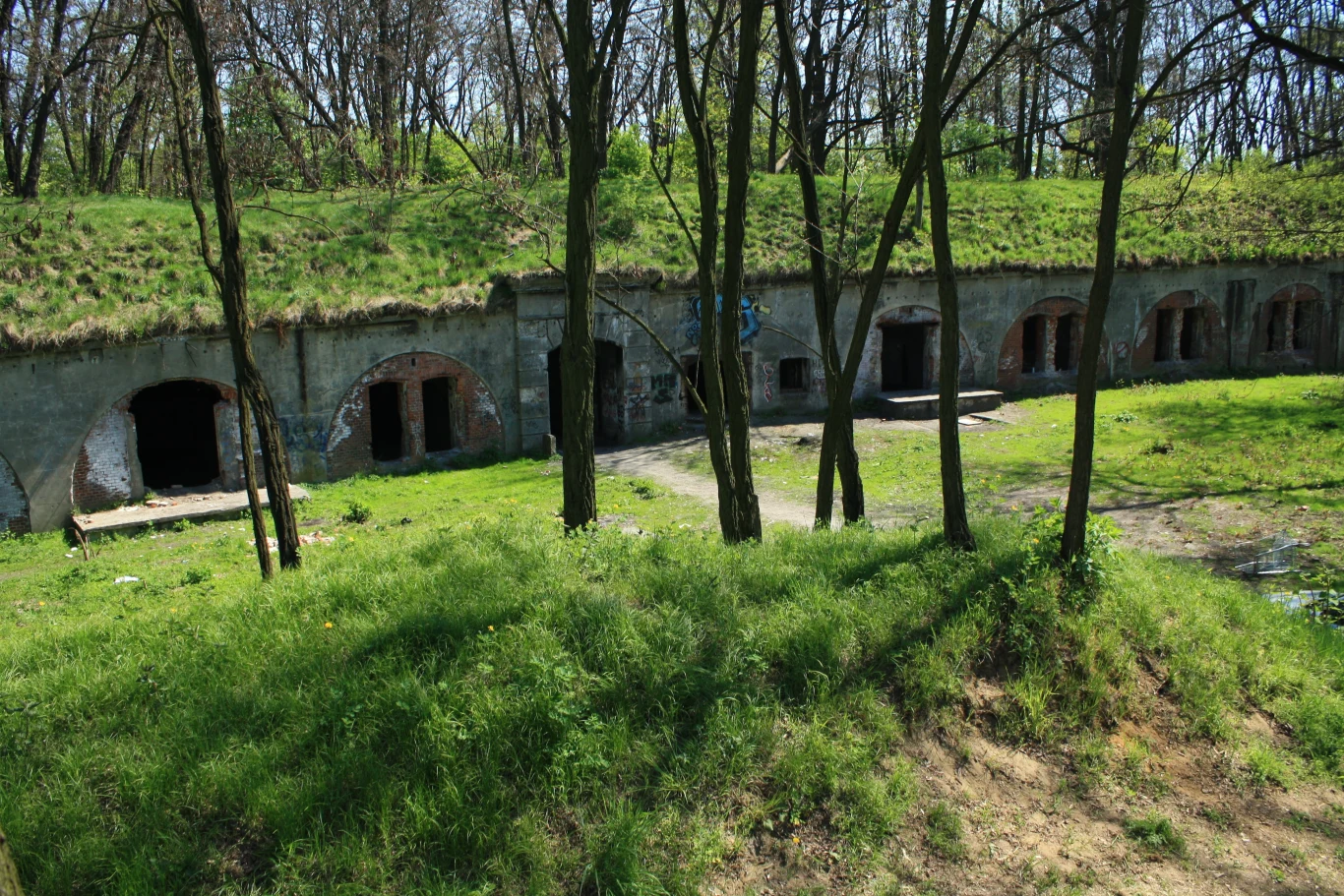 Fort Prokocim w Krakowie Stare ceglane fortyfikacje z łukowatymi wejściami, ukryte w ziemnych nasypach i porośnięte trawą, w tle rosną drzewa bez liści i wczesnowiosenna roślinność.