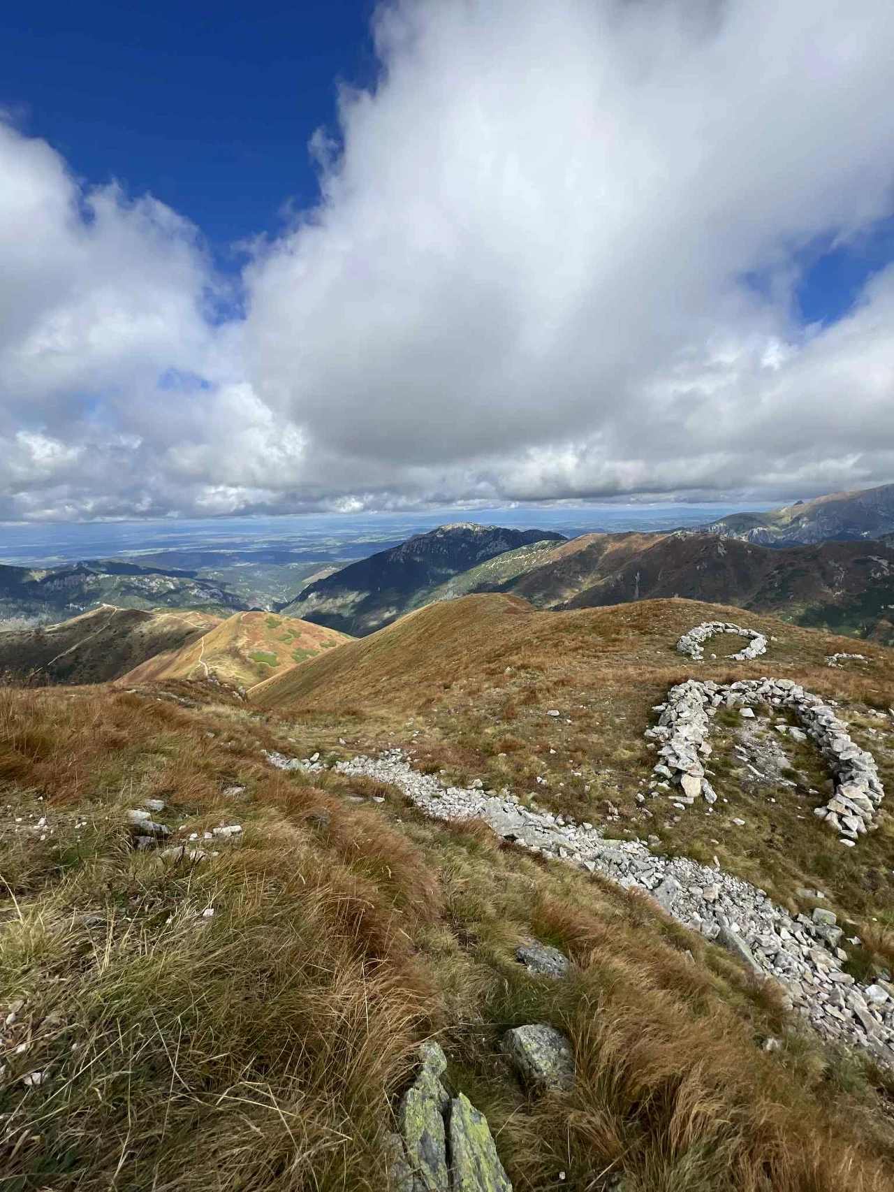 Górski krajobraz z kamienistym szlakiem i figurą człowieka ułożoną z białych kamieni na zboczu, w tle rozciągające się pasma górskie, rozproszone chmury oraz błękitne niebo.