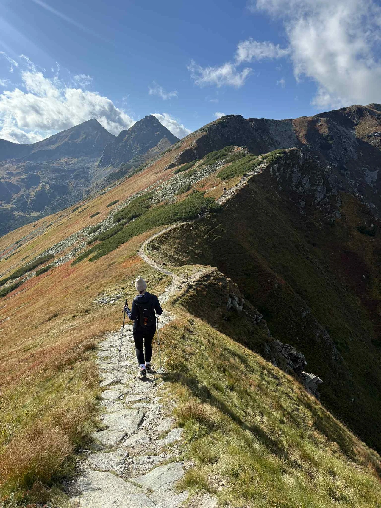 Turysta wędruje kamienistą ścieżką grzbietem górskim w otoczeniu malowniczych wysokich szczytów oraz rozległych połaci trawy pod błękitnym niebem z pojedynczymi chmurami.