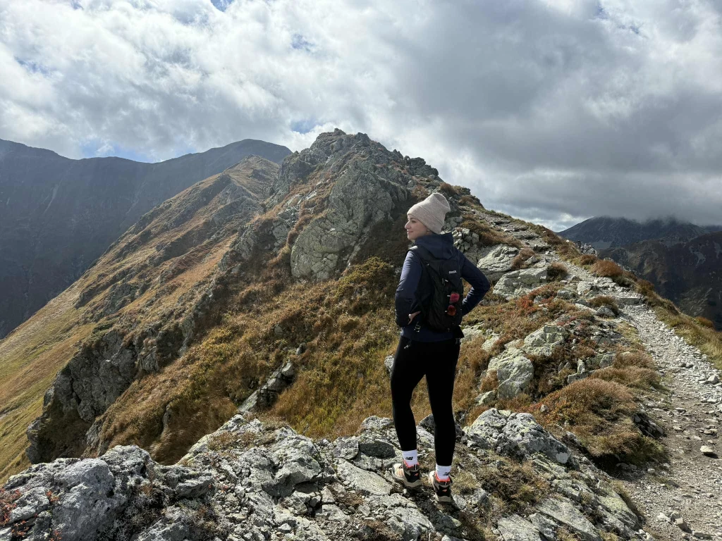 Turystka w sportowym ubraniu stoi na skalistym grzbiecie górskim, spoglądając na otaczający ją krajobraz górski pod zachmurzonym niebem, na tle surowych szczytów i ścieżki prowadzącej w dalsze partie gór.