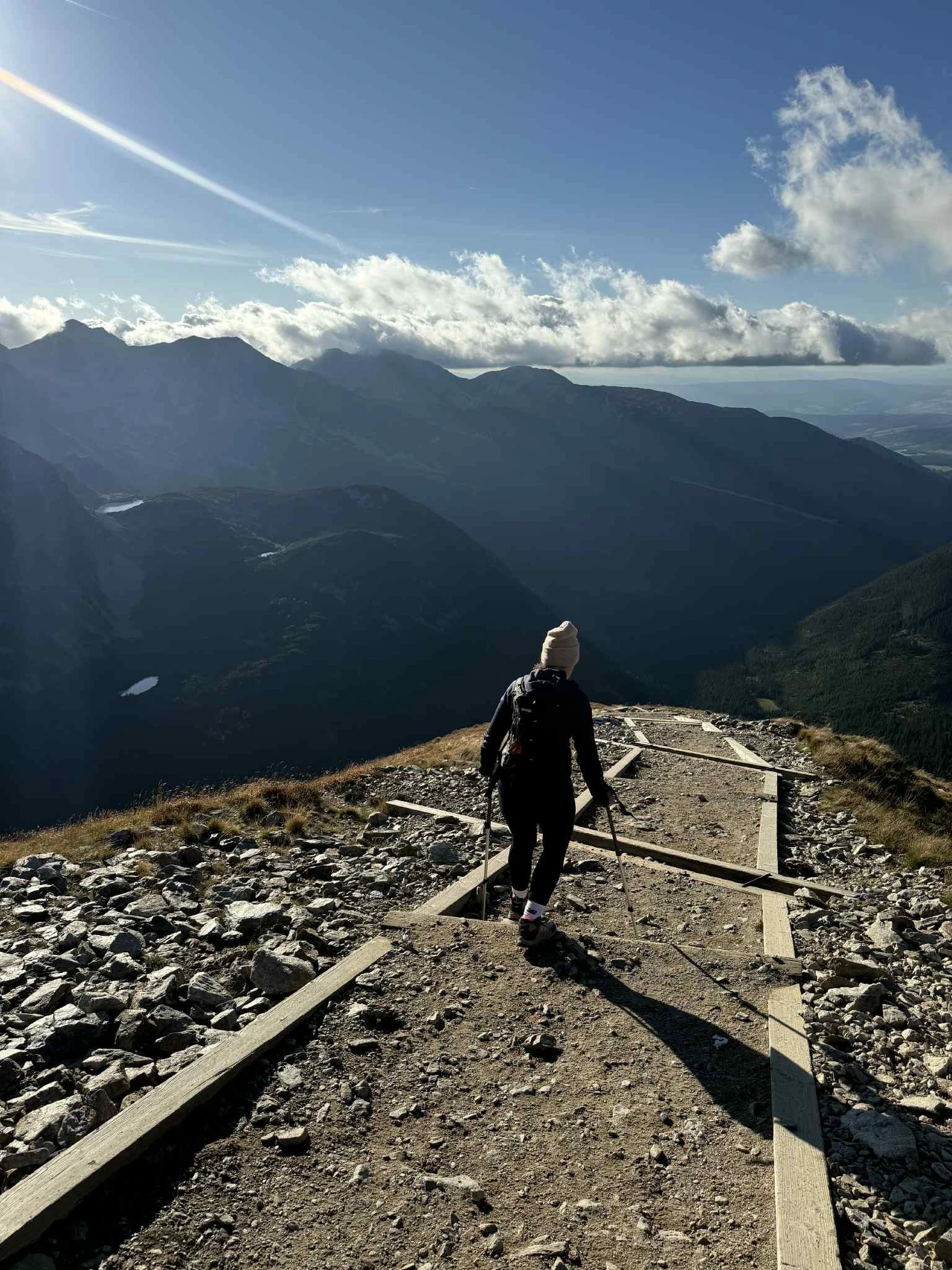 Osoba idąca górskim szlakiem pośród skalistego krajobrazu z rozległym widokiem na górskie szczyty oraz doliny, w oddali rozpościera się panoramiczny widok podświetlony promieniami słońca oraz chmurami.