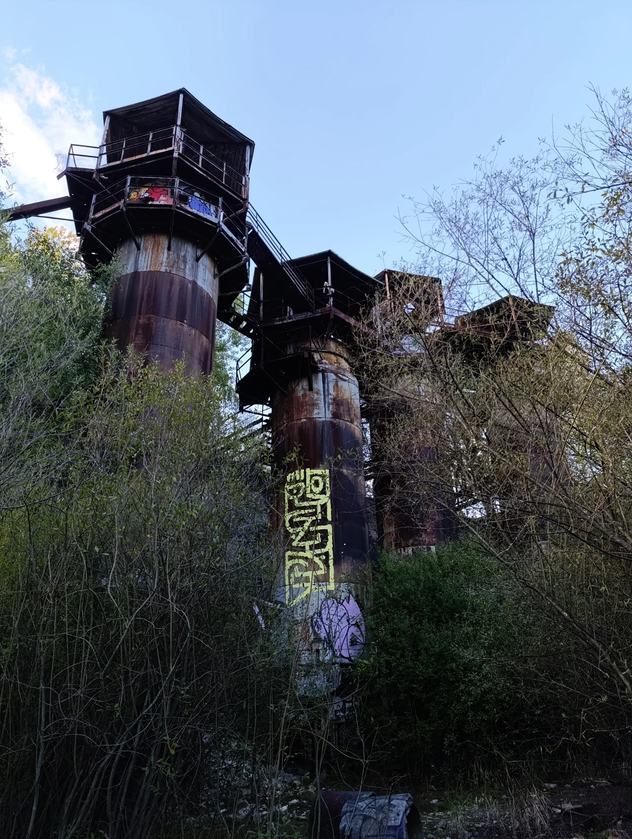 Piece w kamieniołomie Liban w Krakowie Stare, przemysłowe konstrukcje rurowe o znacznych rozmiarach obrastają roślinnością, na jednej z nich widoczne graffiti, a całość zanurzona jest wśród drzew i krzewów.