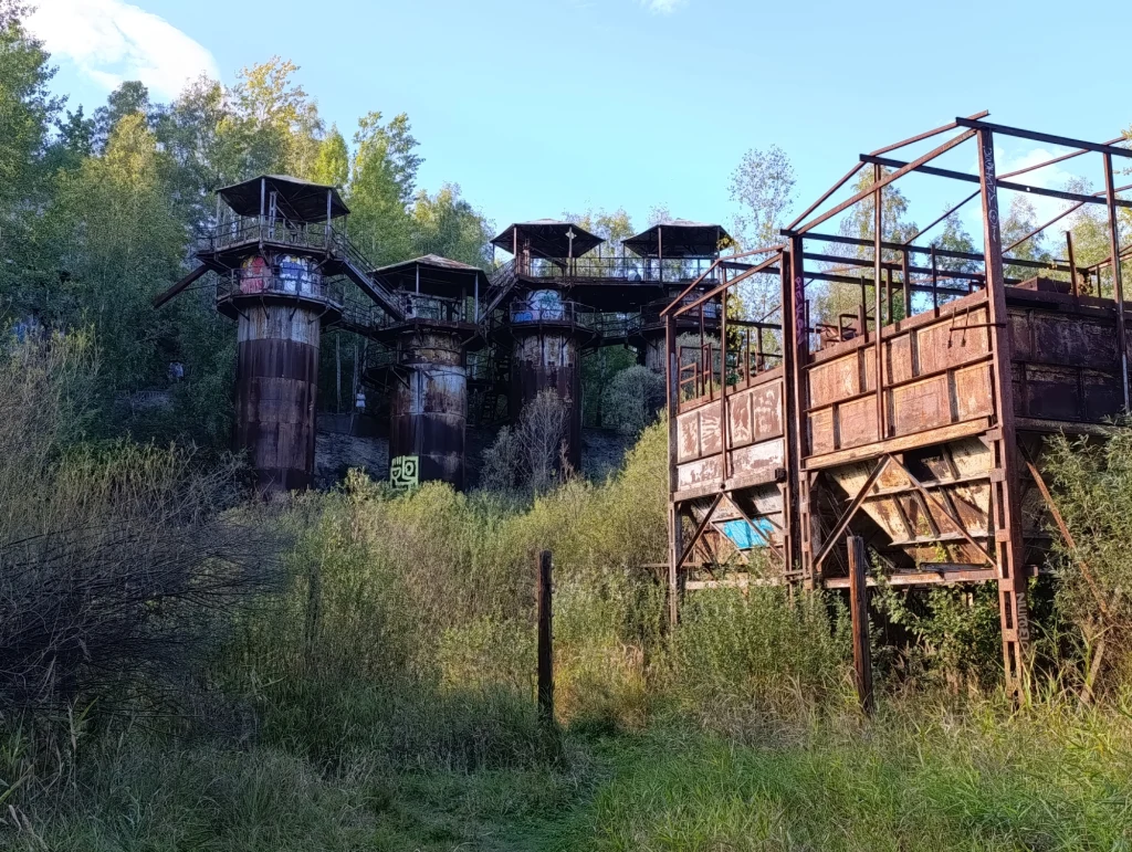 Kamieniołom Liban w Krakowie Opuszczone, zardzewiałe konstrukcje przemysłowe otoczone gęstą roślinnością i drzewami, pozostałości dawnego zakładu, które wtapiają się w naturalny krajobraz.