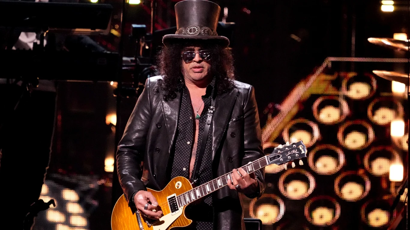 Slash Mężczyzna w charakterystycznym cylindrze i skórzanej kurtce gra na gitarze elektrycznej na scenie koncertowej przy wielobarwnym oświetleniu.