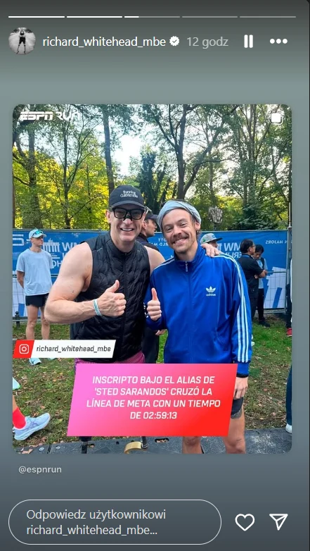 Dwóch mężczyzn po udanym biegu przy trasie maratonu, jeden w sportowej kamizelce i czapce na głowie, drugi w niebieskim dresie sportowym oraz opasce, obaj uśmiechnięci pokazują gest kciuka w górę, w tle drzewa i inni uczestnicy biegu