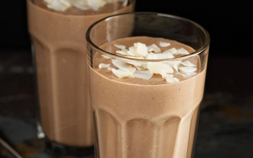 Jak przyrządzić kawę z mleczkiem kokosowym? Szklanka gęstego koktajlu czekoladowego udekorowanego wiórkami kokosowymi, w tle widoczna jest druga szklanka z tym samym napojem.