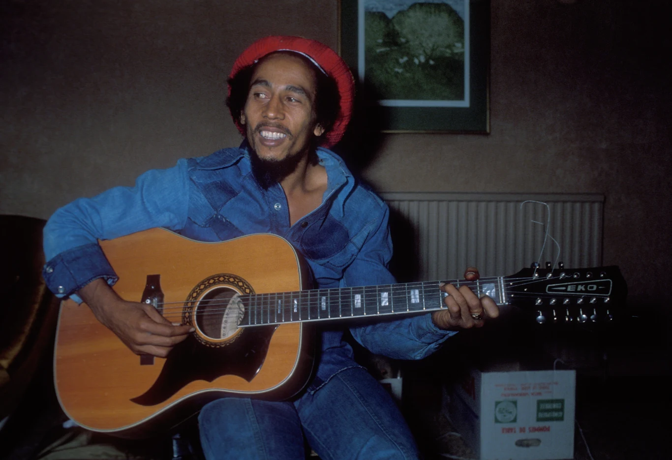 Bob Marley Uśmiechnięty mężczyzna w dżinsowej kurtce i czerwonej czapce gra na dwunastostrunowej gitarze akustycznej, siedząc w przytulnym, domowym wnętrzu.