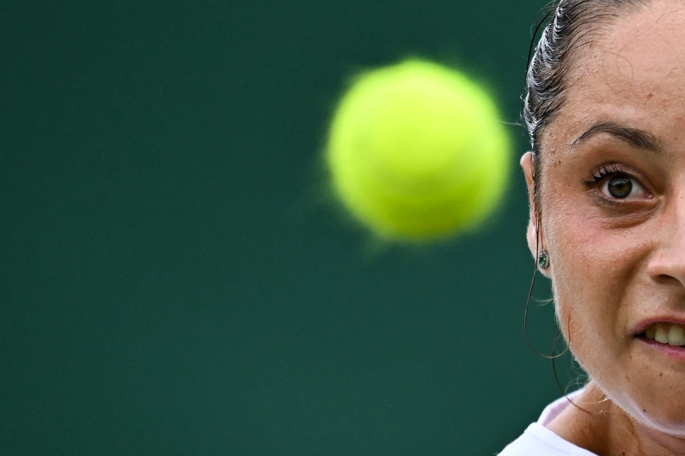 Ekspresyjna twarz tenisistki koncentrującej się podczas gry, z dynamicznie rozmytą piłką tenisową unoszącą się na pierwszym planie na tle zielonego kortu.