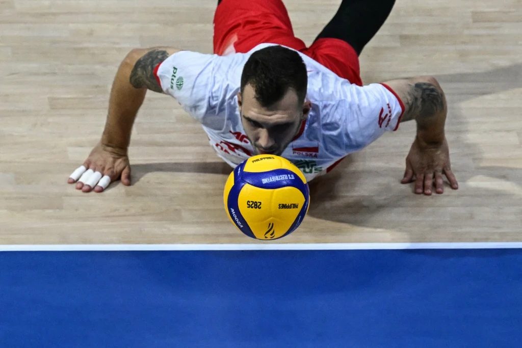 Norbert Huber Zawodnik siatkarski w białej koszulce i czerwonych spodenkach podczas dynamicznej akcji tuż przy parkiecie, skupiony na piłce siatkowej unoszącej się blisko jego twarzy.