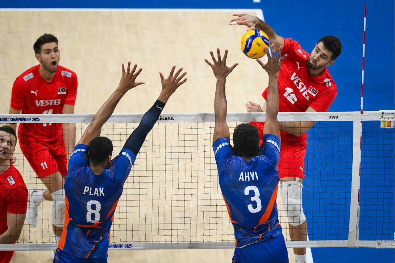Adis Lagumdzija (w ataku) Sportowa rywalizacja drużyn siatkarskich, zawodnik w czerwonym stroju atakuje piłkę nad siatką, dwóch zawodników w niebieskich strojach próbuje zablokować atak, widoczny dynamiczny moment gry.