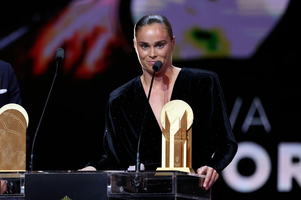 Ewa Pajor odebrała nagrodę Gerda Muellera Kobieta w eleganckiej czarnej sukni stoi przy mównicy z mikrofonami, trzymając złotą statuetkę na tle rozmytego, kolorowego ekranu.