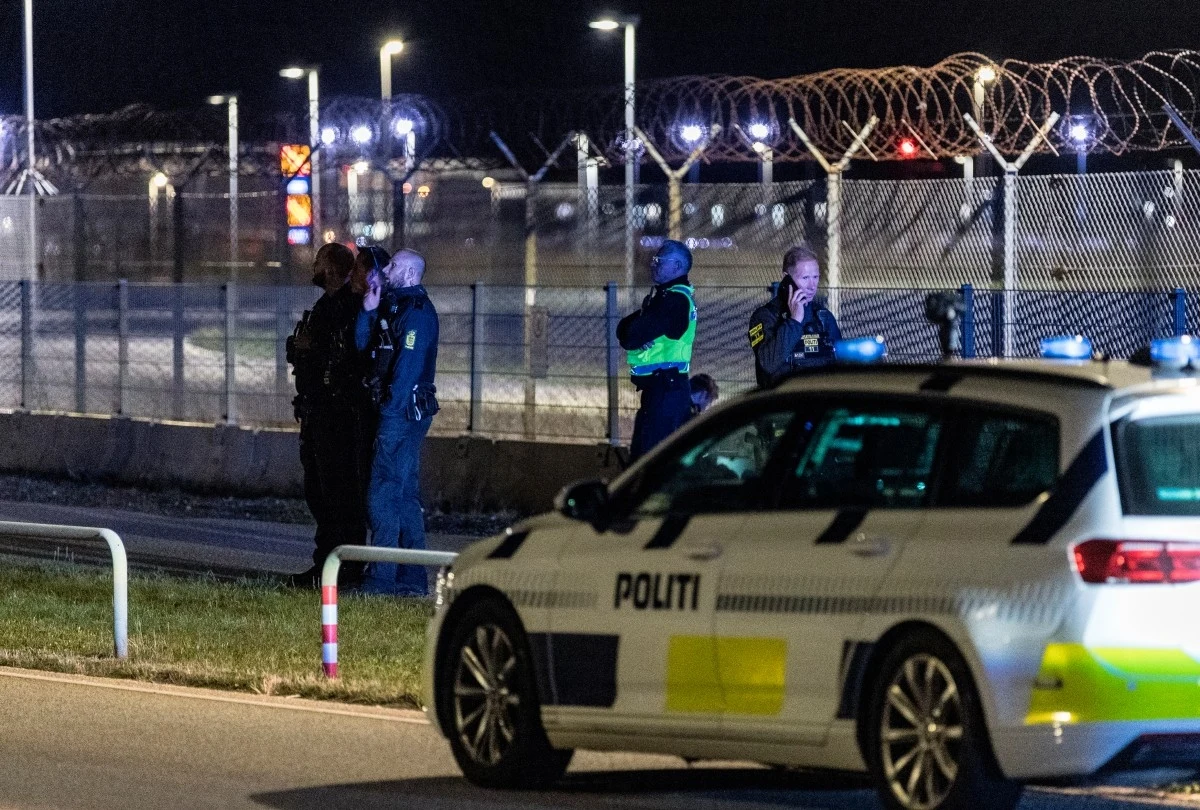 W związku z incydentem na lotnisku w Kopenhadze pojawiła się policja Policjanci rozmawiają przy ogrodzeniu z drutem kolczastym, na pierwszym planie radiowóz policyjny z napisem 'POLITI', w tle oświetlenie i zabezpieczenia terenu.