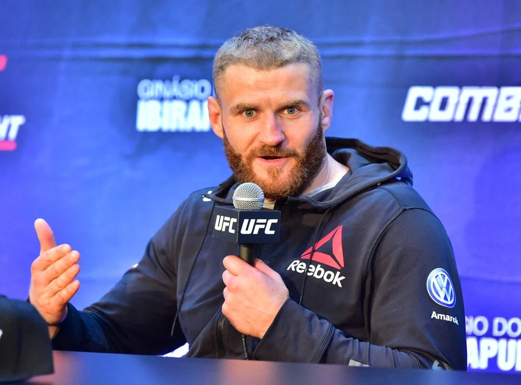 Mężczyzna z brodą w sportowej bluzie z logotypami UFC, Reebok i Volkswagena siedzi przy stole z mikrofonem w ręce i gestykuluje, za nim widoczna ściana z logotypami sponsorów.