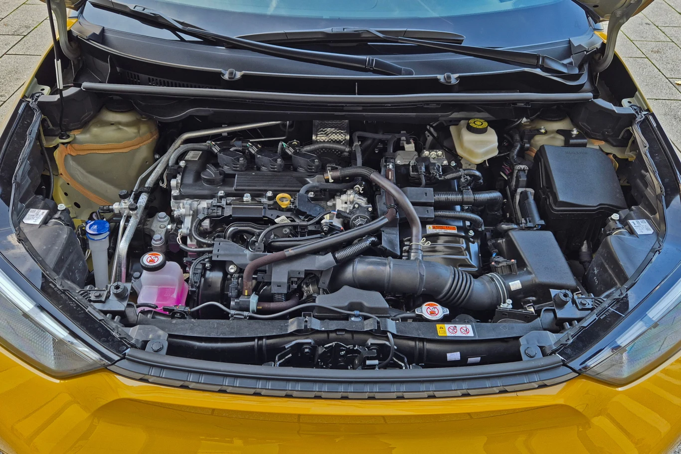 Komora silnika nowoczesnego samochodu osobowego z widocznymi elementami układu chłodzenia, przewodami, zbiornikiem płynu chłodniczego i akumulatorem, obudowana z przodu żółtym nadwoziem pojazdu.