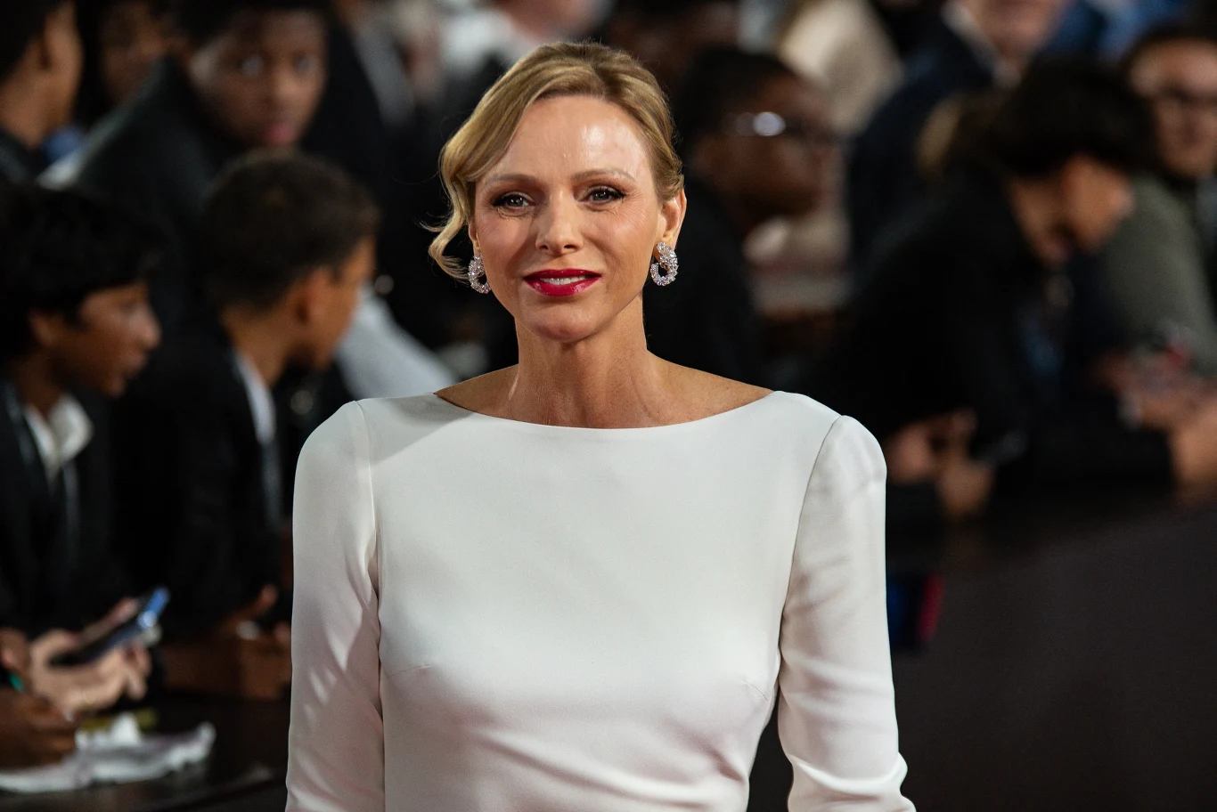 Elegancka kobieta w białej sukni z perłowymi kolczykami uśmiecha się, stojąc na tle rozmytego tłumu podczas oficjalnego wydarzenia.