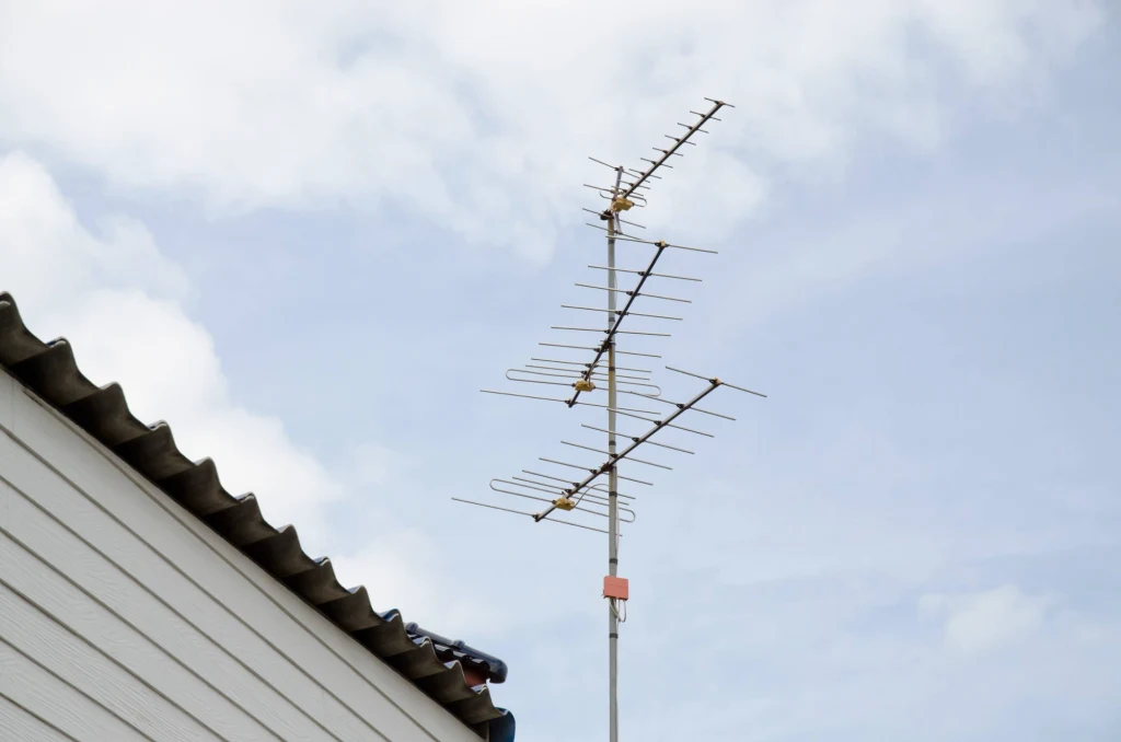 Metalowa antena telewizyjna zamontowana na dachu budynku z falistą, szarą blachą na tle jasnego, lekko zachmurzonego nieba.