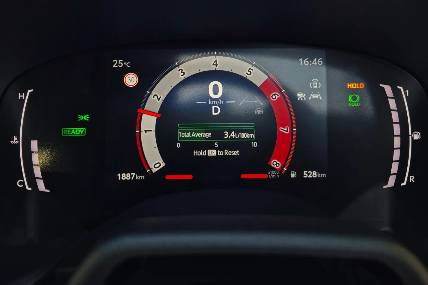 Cyfrowy panel wskaźników w samochodzie z prędkościomierzem centralnym, wskaźnikiem poziomu paliwa oraz informacjami o przebiegu i średnim spalaniu, otoczony ikonami sygnalizacyjnymi.