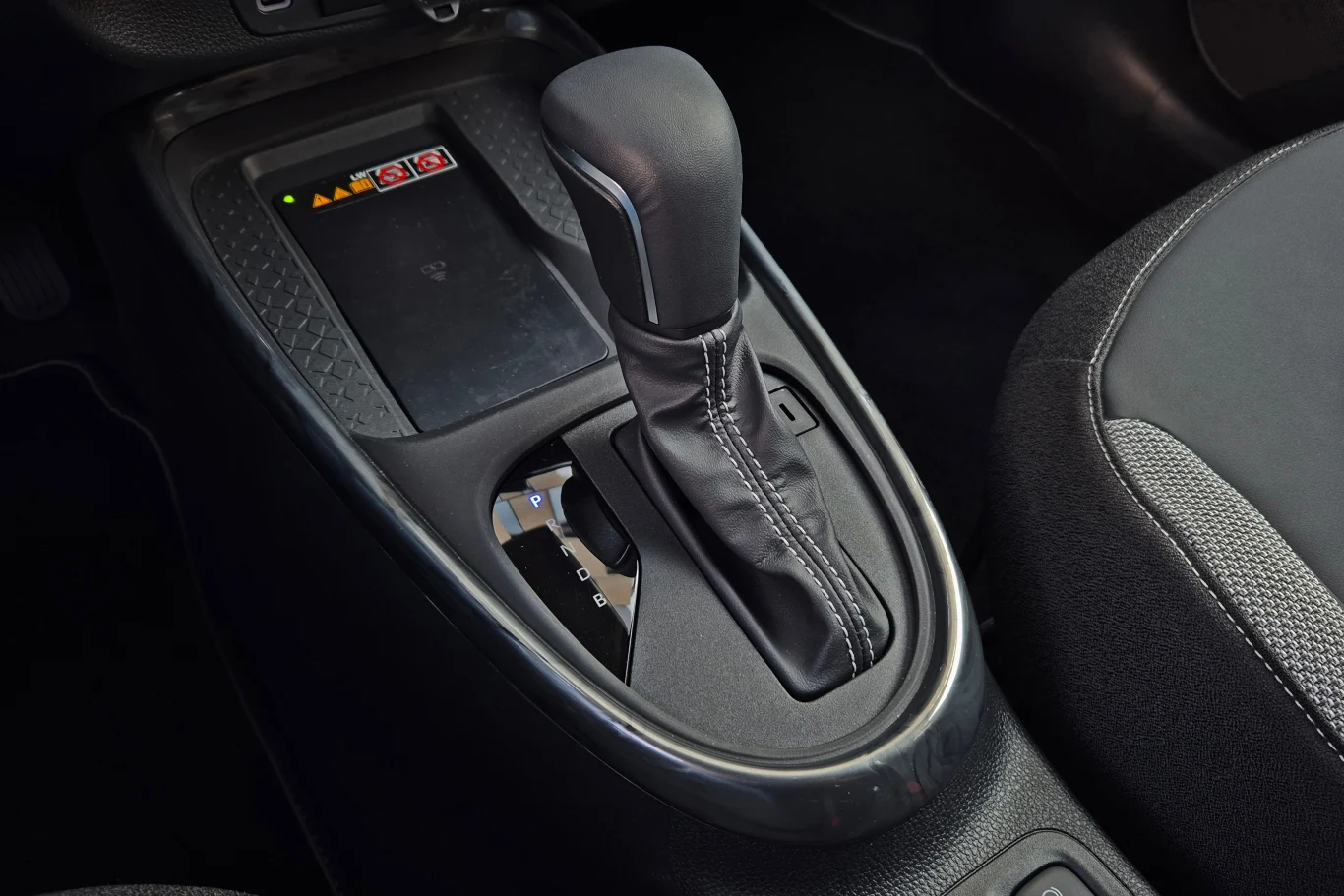 Dźwignia automatycznej skrzyni biegów wraz z otaczającym ją panelem środkowym samochodu, widoczne detale pokrycia fotela oraz wykończenia wnętrza auta.