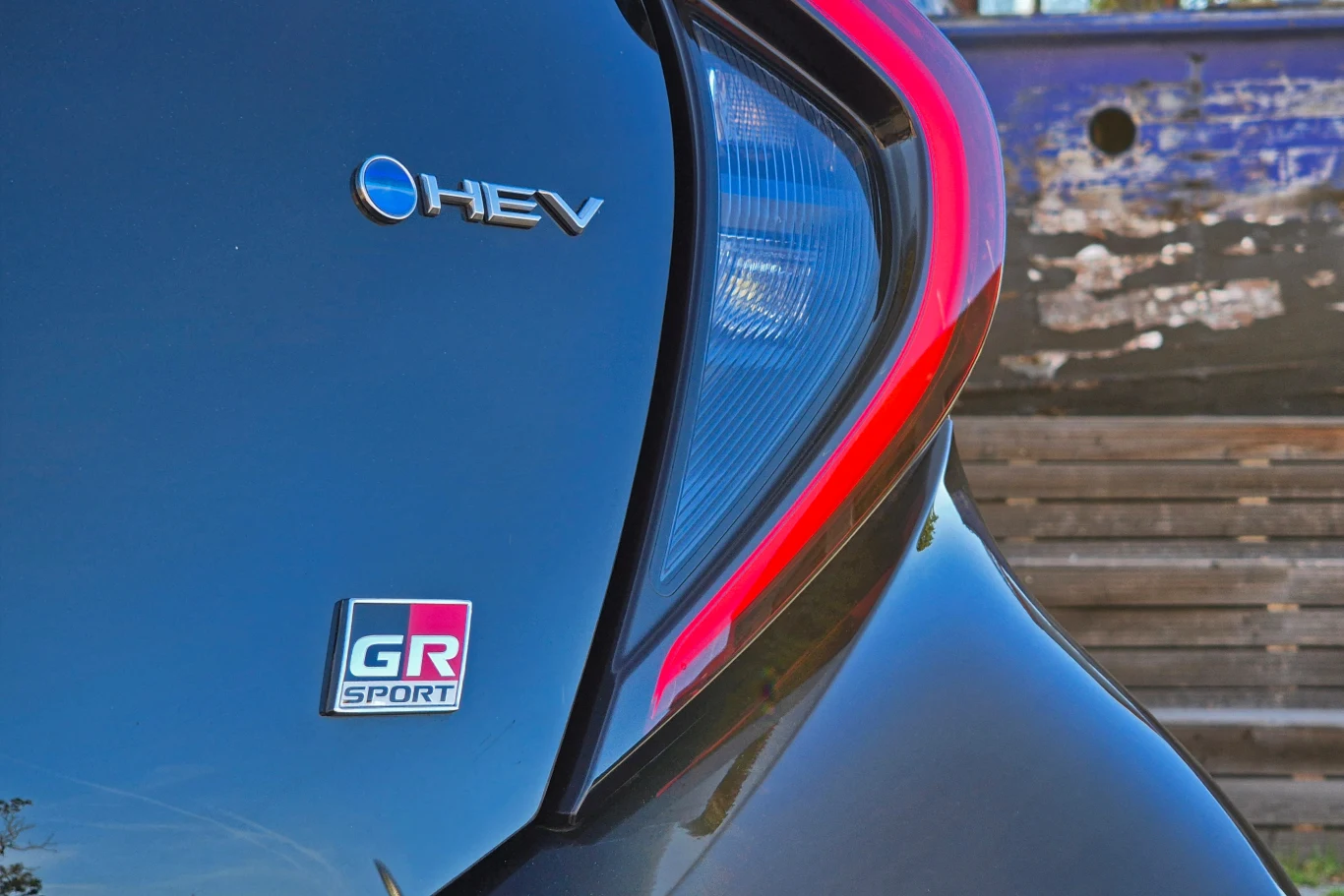 Tylna część czarnego samochodu z widoczną plakietką GR SPORT oraz oznaczeniem HEV, podkreślona nowoczesnym tylnym światłem i refleksami na lakierze, w tle rozmazana drewniana powierzchnia.