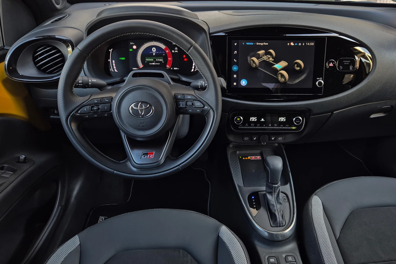 Nowoczesna deska rozdzielcza samochodu Toyota z kierownicą, cyfrowym wyświetlaczem oraz dużym ekranem dotykowym zintegrowanym z centralnym panelem; widoczne fotele pokryte materiałem i elementy kokpitu.