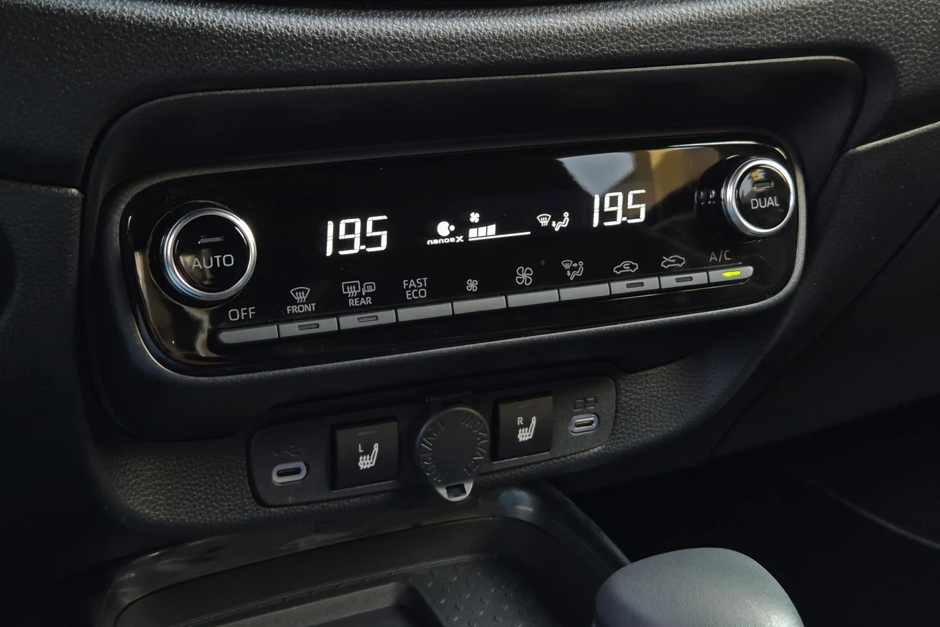 Panel sterowania klimatyzacją samochodową z cyfrowym wyświetlaczem wskazującym temperaturę 19,5°C po obu stronach, przyciskami do regulacji nawiewu, ogrzewania, klimatyzacji oraz gniazdem zasilania poniżej.