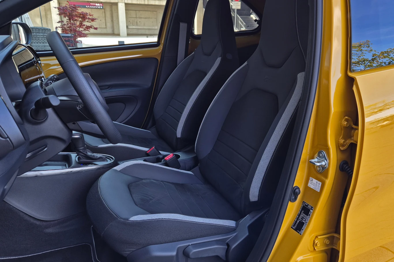 Przednie siedzenia samochodu z czarną tapicerką, kierownica, deska rozdzielcza i fragment żółtych drzwi, wnętrze pojazdu ukazane z otwartymi drzwiami po stronie kierowcy