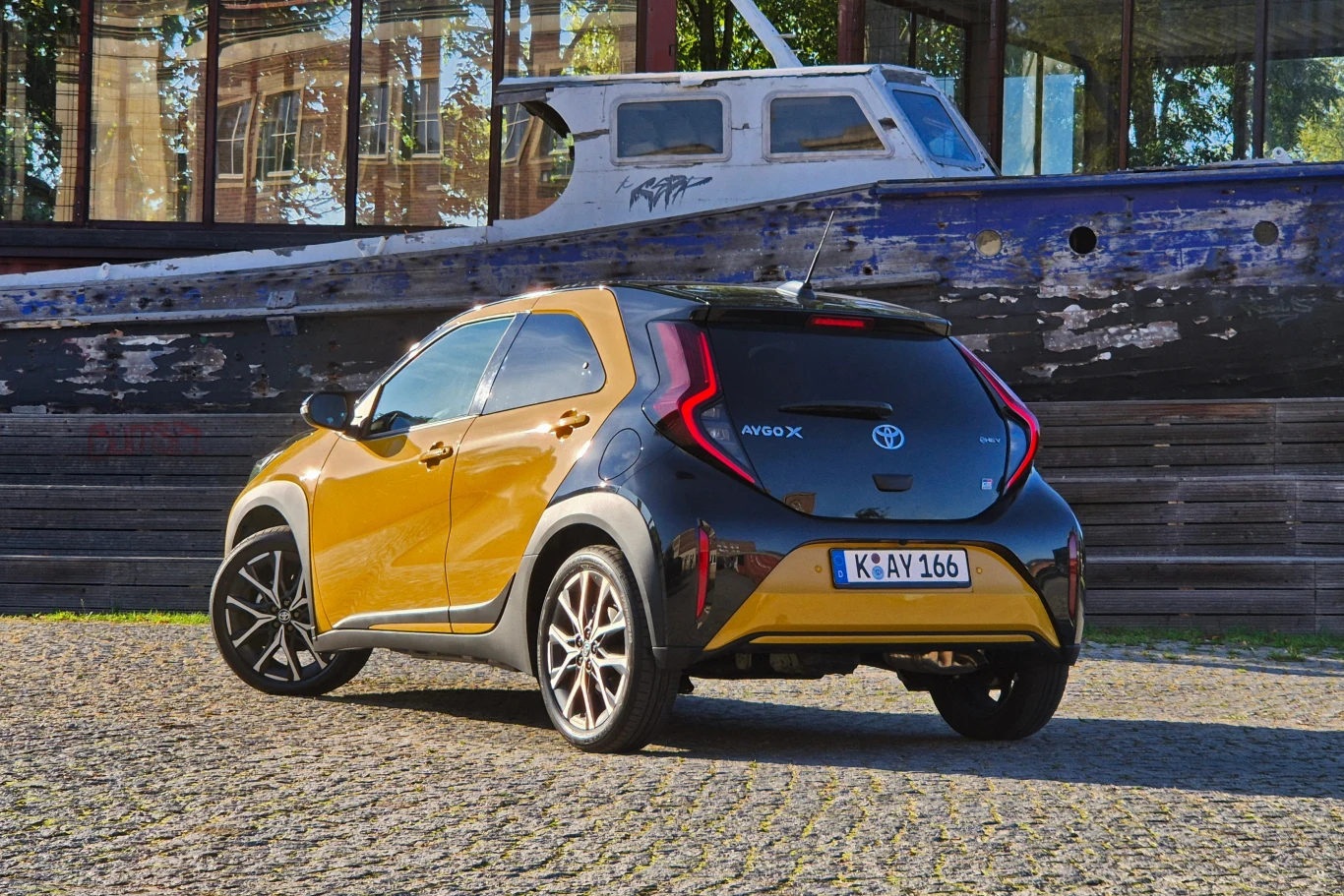 Nowoczesny, żółto-czarny samochód marki Toyota Aygo X stoi zaparkowany na brukowanej nawierzchni, w tle widoczny stary, zniszczony kadłub łodzi ustawionej na suchym lądzie.