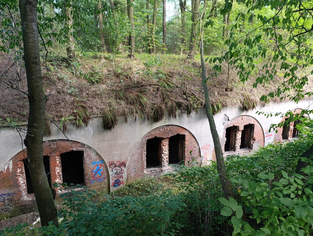 Fort 50 Prokocim Opuszczona, stara fortyfikacja ukryta wśród gęstej zieleni, z widocznymi wejściami do wnętrza i śladami graffiti na ceglanych ścianach, na dachu rośnie trawa i drobne krzewy.