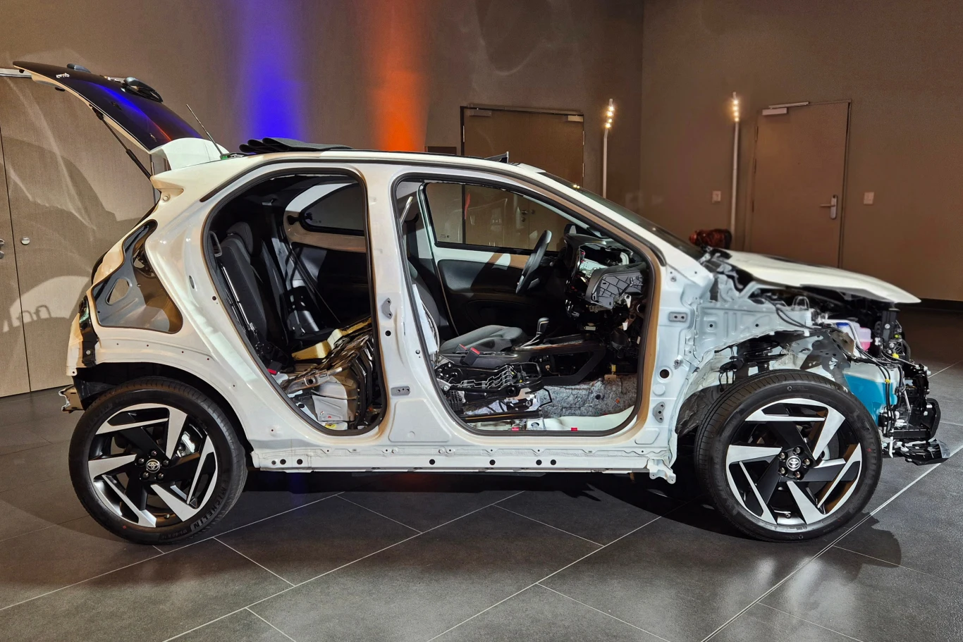 Samochód osobowy w białym kolorze, pozbawiony większości elementów karoserii oraz wnętrza, ukazujący wnętrze konstrukcji i podzespoły mechaniczne; drzwi oraz maska zdemontowane, widoczne detale techniczne auta.
