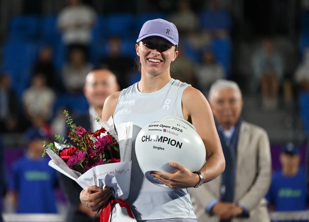 Iga Świątek z trofeum Korea Open Sportowa zawodniczka w jasnej czapce i koszulce trzyma bukiet czerwonych i różowych kwiatów oraz dużą białą piłkę z napisem Champion, w tle kilka osób i widownia.