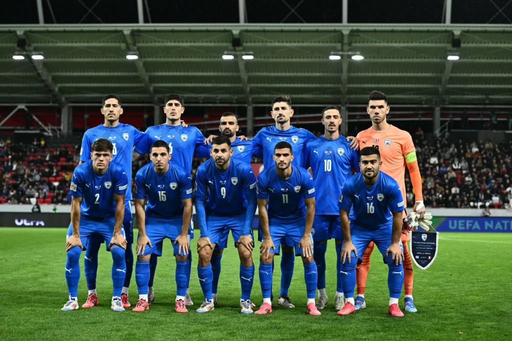 Reprezentacja Izraela Piłkarska drużyna w niebieskich strojach ustawiona w dwóch rzędach na murawie stadionu, za nimi trybuny pełne kibiców oraz nowoczesna konstrukcja dachu.