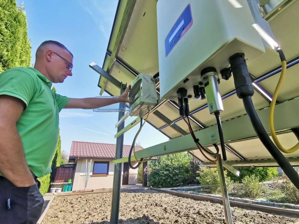 Czy prąd z fotowoltaiki jest wykorzystywany na bieżąco? Kiedy dochodzi do strat? Mężczyzna w zielonej koszulce sprawdza instalację elektryczną pod panelem fotowoltaicznym zamontowanym na zewnątrz, na tle ogrodu i małego budynku.
