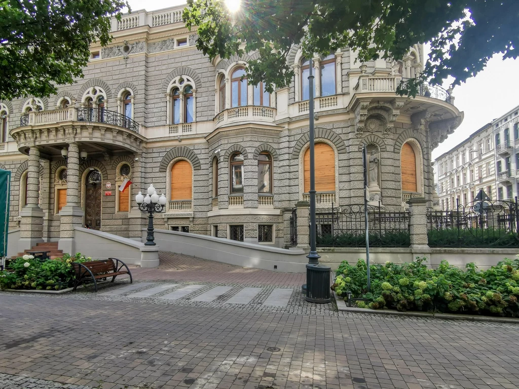 Zabytkowy budynek o bogato zdobionej elewacji, okna zabezpieczone deskami, na tle kamiennego chodnika i zieleni. Flagę narodową umieszczono przy wejściu. Otoczenie zadbane, bez ludzi.