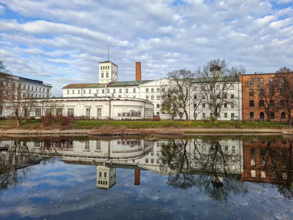 Historyczny, biały budynek z charakterystyczną wieżą i czerwonym kominem, otoczony drzewami, odbija się w spokojnej tafli wody w jesiennej scenerii.