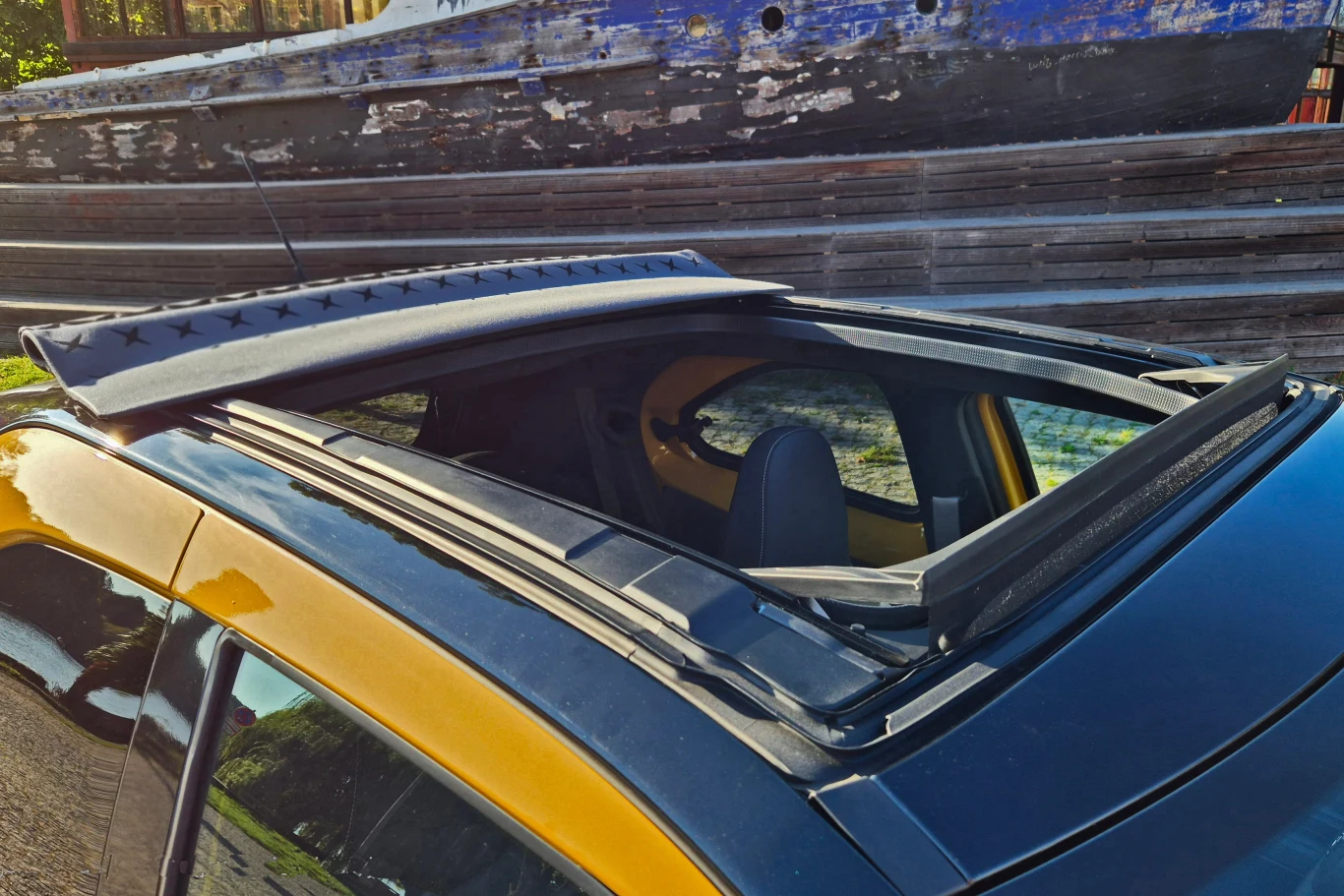 Otwarty dach panoramiczny samochodu o żółtym lakierze, w tle widoczna drewniana łódź o zużytym wyglądzie.