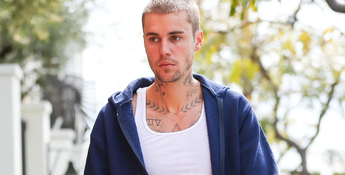 Justin Bieber Młody mężczyzna o krótkich włosach i widocznych tatuażach na klatce piersiowej ubrany w biały podkoszulek oraz niebieską rozpinaną bluzę, stoi na tle zieleni i nieostrego tła ulicznego.