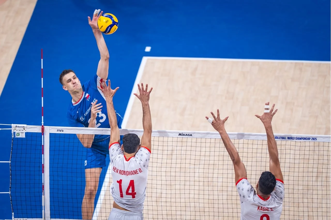 Silny atak siatkarza w niebieskim stroju, wykonującego skok z zamiarem zbicia piłki, którą próbuje zablokować dwóch zawodników drużyny przeciwnej ubranych w białe stroje z czerwonymi akcentami.