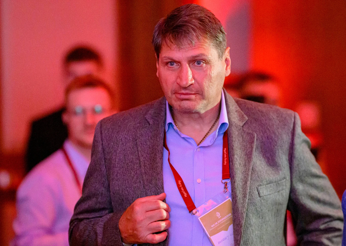 Andrzej Gołota Mężczyzna w marynarce i niebieskiej koszuli z czerwoną smyczą konferencyjną na szyi stoi na tle rozmytych postaci i czerwonego światła, skupiony patrzy przed siebie, trzymając dłoń przy klatce piersiowej.