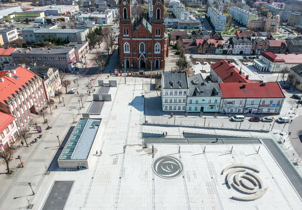 Rozległy, jasny plac miejski z geometrycznymi wzorami na płycie, otoczony budynkami – w tym strzelistym kościołem z czerwonej cegły oraz kamienicami i obiektami usługowymi; widok z lotu ptaka.