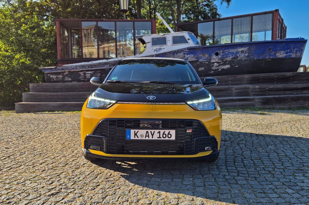 Żółto-czarny samochód marki Toyota ustawiony na brukowanym placu, w tle widoczny stary, niebiesko-biały kuter rybacki oraz nowoczesny budynek z dużymi przeszkleniami, całość otoczona zielenią.
