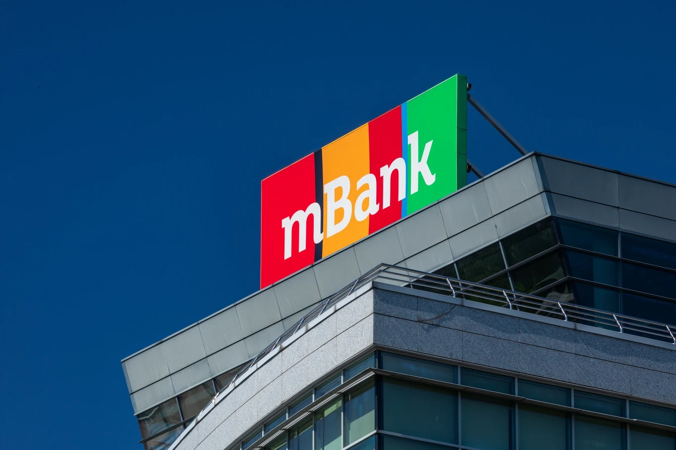 mBank zostanie przejęty? Eksperci oceniają wyniki giełdowe Kolorowe logo mBanku na szczycie nowoczesnego budynku, widoczne na tle jasnego, niebieskiego nieba.