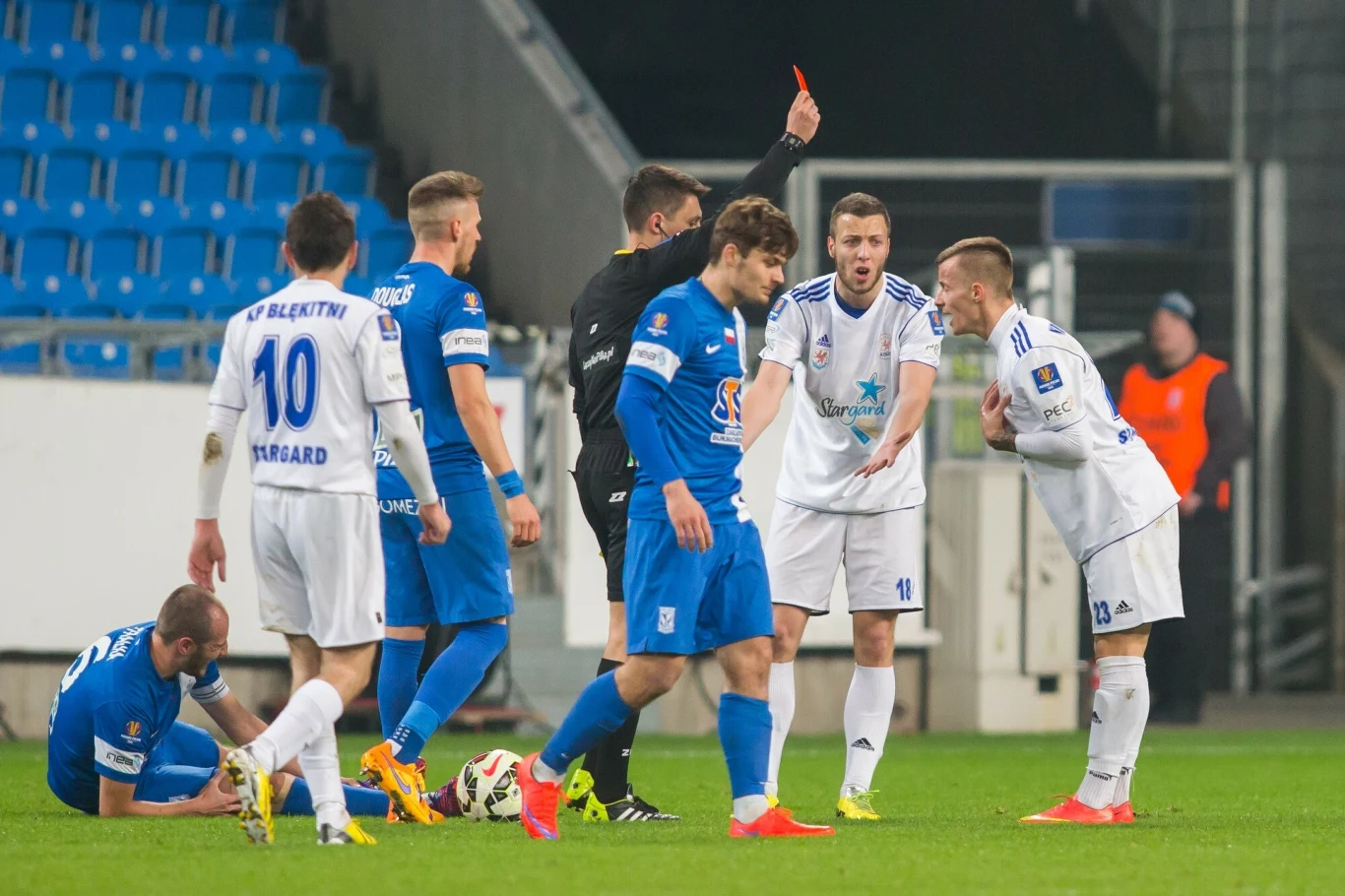 Sędzia pokazuje czerwoną kartkę zawodnikowi podczas meczu piłki nożnej, wokół obecni są piłkarze obu drużyn, jeden z zawodników siedzi na murawie trzymając nogę, a reszta żywiołowo reaguje na decyzję sędziego.